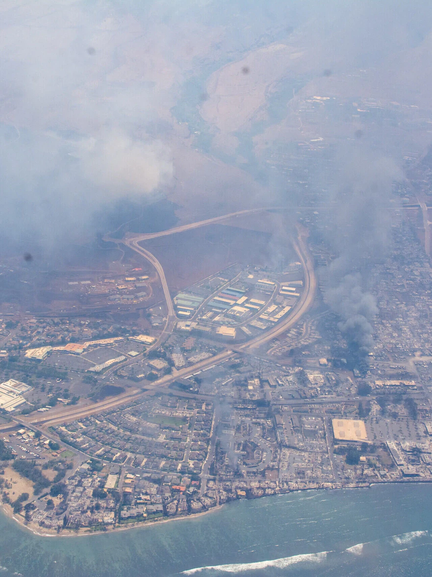 An aerial view of Lahaina under smoke, the burned town beside the shore.