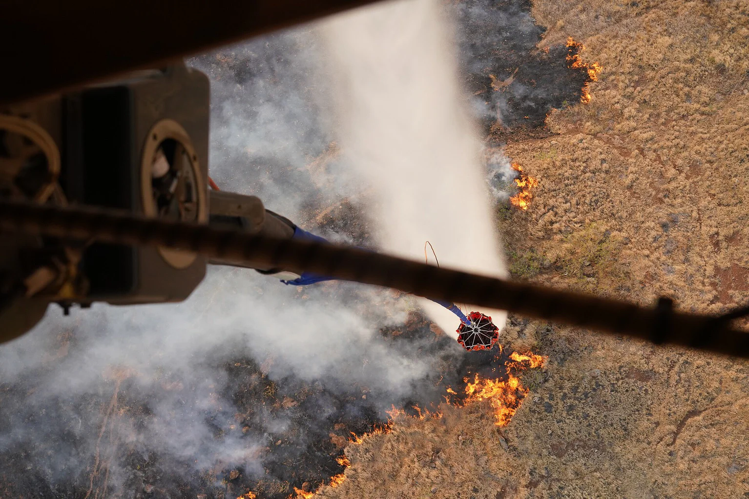 A helicopter’s water bucket drops a plume of water over a burning hillside.