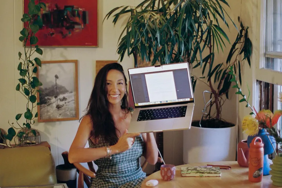 Kenna Reed smiles at a table in a plant-filled room, holding a laptop up beside her.
