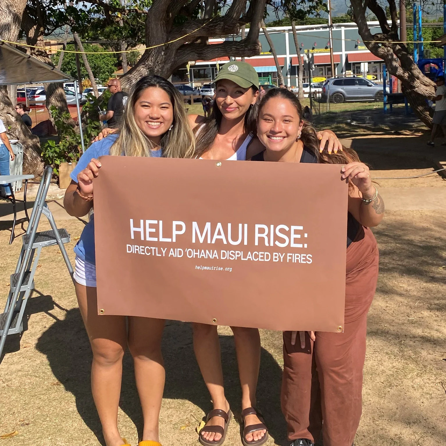 Three volunteers hold a banner reading “Help Maui Rise: Directly aid ‘Ohana displaced by fires” under a tree in a park.
